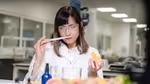 A woman in a lab coat and safety googles smells a paper-smelling strip while holding an orange in her other hand.