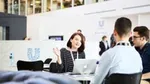 A group of people in a modern office engaged in discussion around a table, with one person gesturing while speaking. An open laptop is on the table, and the Unilever logo and branding is visible on a white wall in the background.